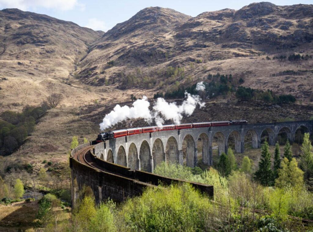 A red steam train crosses a cement curved viaduct with a huge plume of white steam. Rugged mountains form the backdrop. The Jacobite train on Glenfinnan Viaduct.