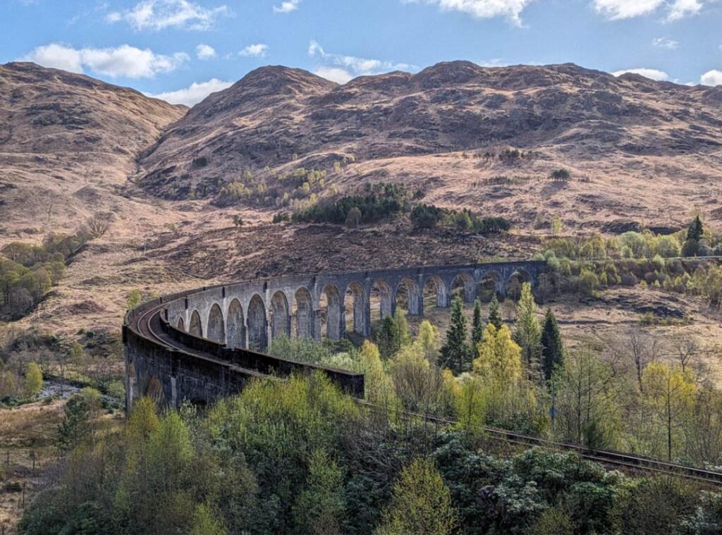 The curved cement arches of Glenfinnan Viaduct with a railway line running over it. Trees dot the foreground and the background is rugged, tawny-coloured and mountainous. 