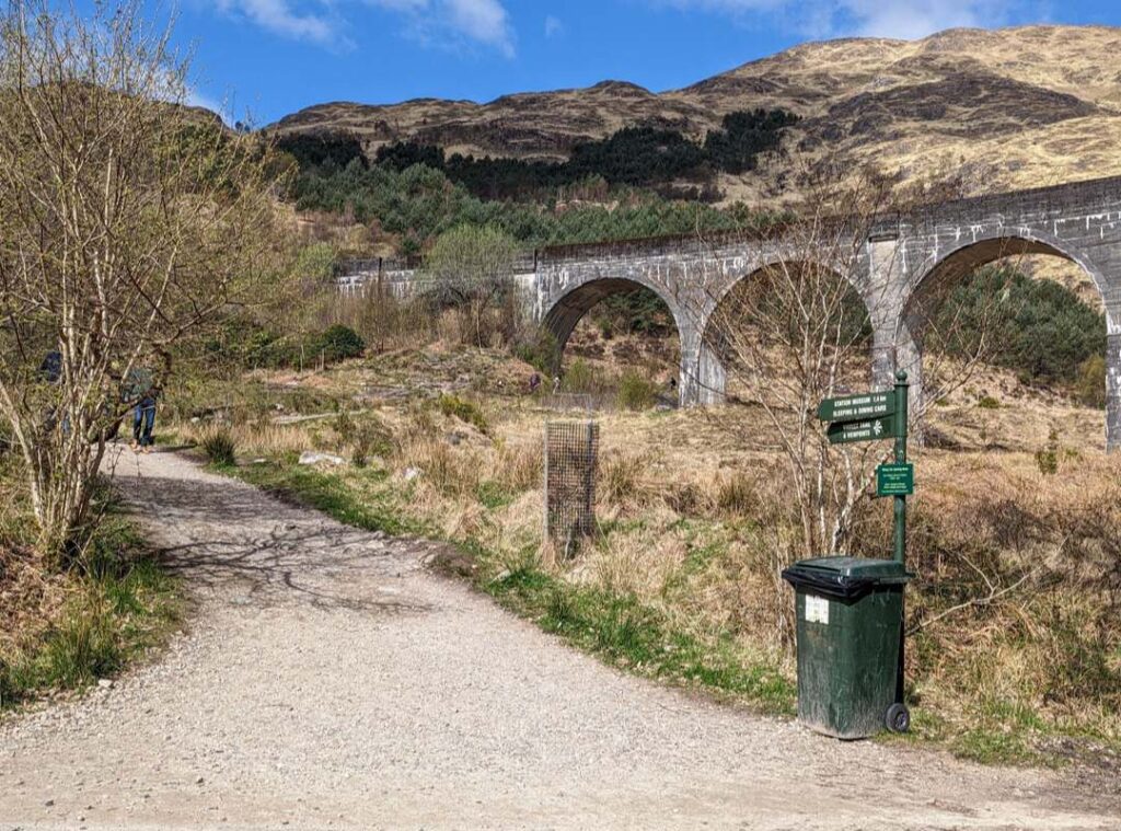 A wide gravel path leads up out of sight. The archways of Glenfinnan Viaduct appear to the right and a green signpost points to the viewpoint. 