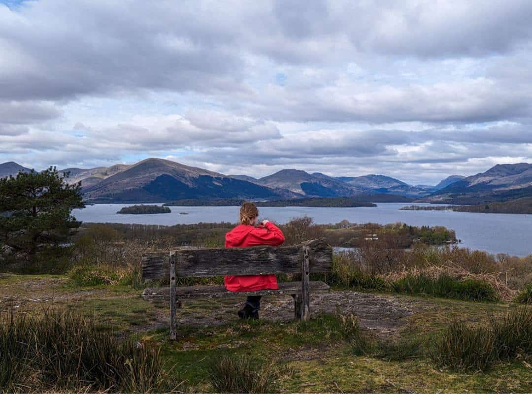 11 Best Day Trips From Glasgow By Public Transport 9 A girl in a red coat sits on a bench on top of a hill looking out across a loch dotted with islands. Hills surround the background. Inchcailloch on Loch Lomond.