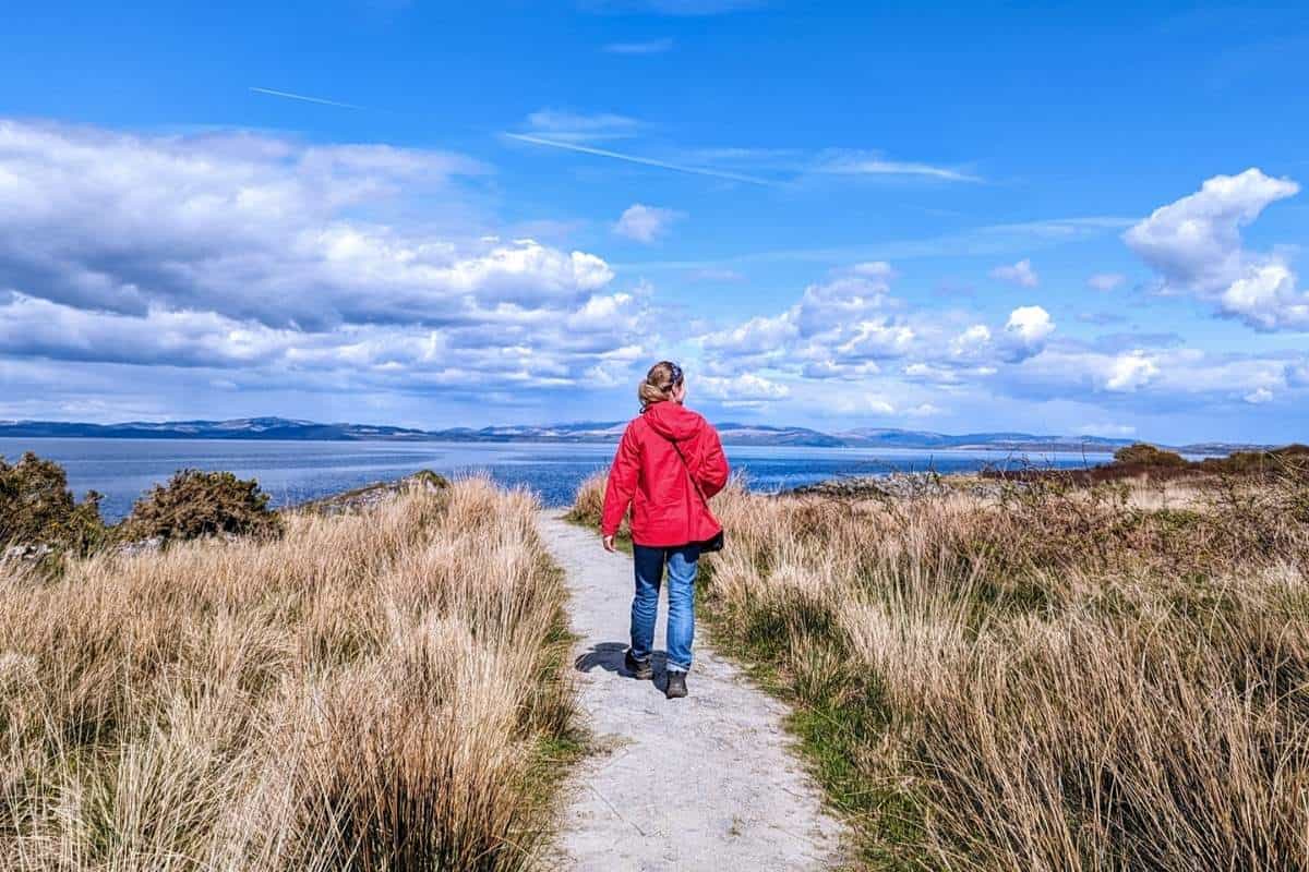 11 Best Day Trips From Glasgow By Public Transport 1 A woman wearing a red jacket and blue jeans walks along a coastal path on the Isle of Arran, one of the best day trips from Glasgow by public tranport.
