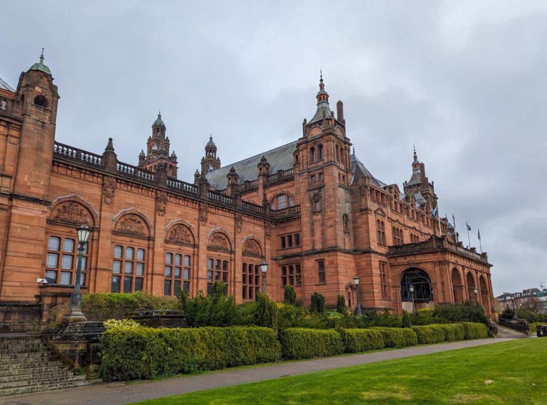 The terracotta coloured building of Kelvingrove Museum with a manicured lawn and hedges on a cloudy day in Glasgow. 
