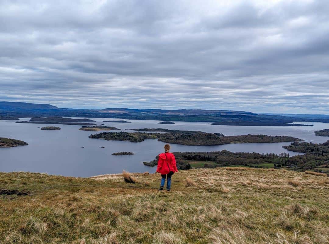 11 Best Day Trips From Glasgow By Public Transport 6 A girl in a red coat stands on a grassy hill and looks down on a loch dotted with islands. There are hills in the far distance. An alternative to Conic Hill, Beinn Dubh in Luss is one of the best day trips from Glasgow by public transport.