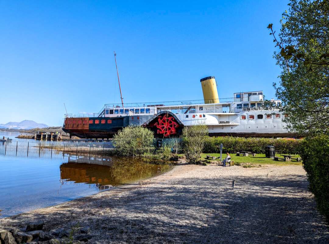 11 Best Day Trips From Glasgow By Public Transport 3 A large white, black and red steamer boat with a yellow chimney is moored on the shores of a loch. There's a beach in the foreground and a view of hills in the background.
