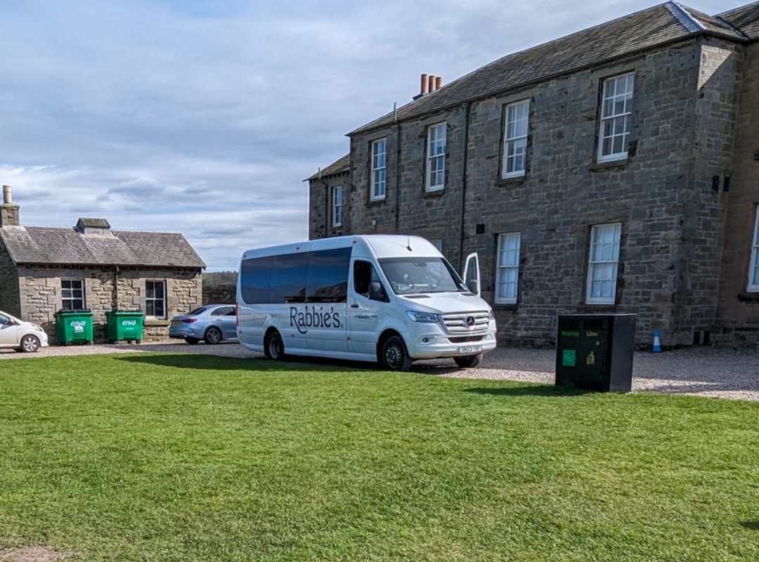 A white minivan with the Rabbie's tours logo is parked outside of a stone building with a patch of grass at the front. 