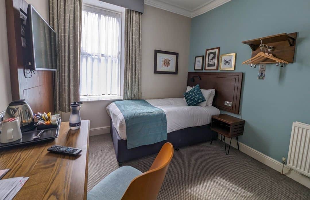 Image of a room with a single bed and green details on it. A head board with pictures behind the bed and there's a desk with a kettle and tea in the left foreground. It's a room at Balloch House Hotel. 
