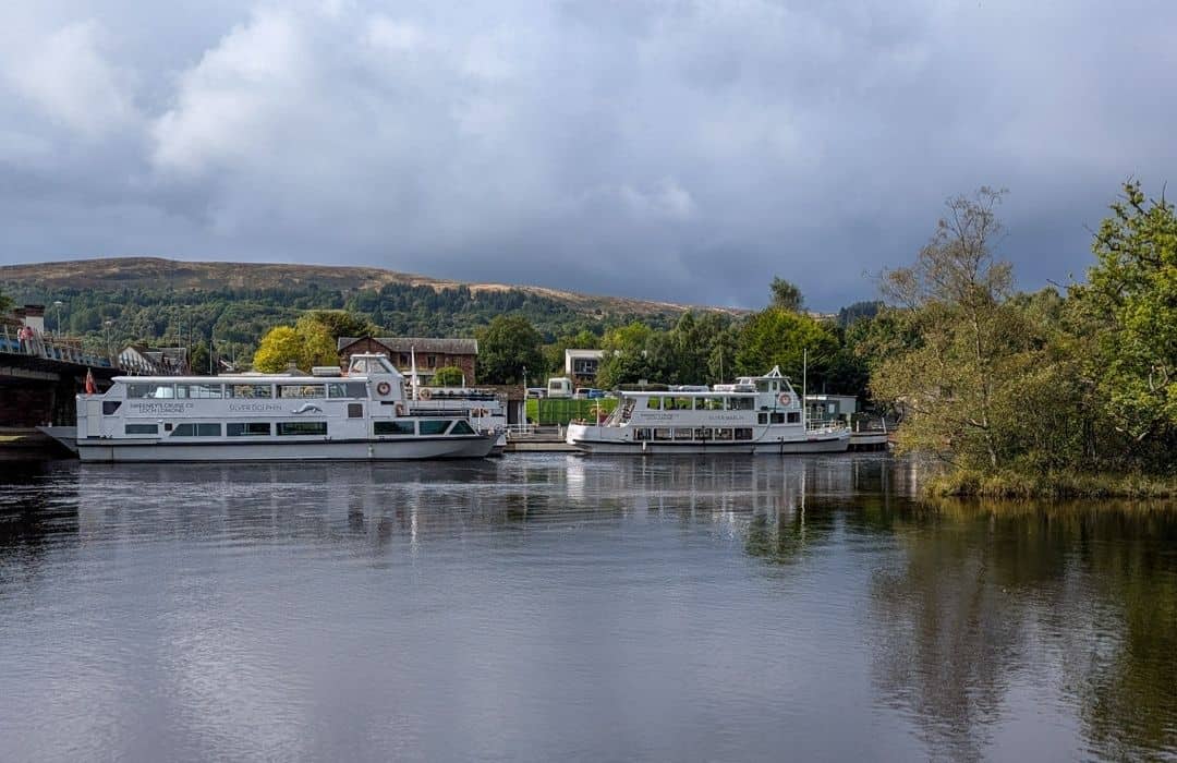 Two white boats are anchored on the calm waters of the River Leven next to some trees. Tree-covered hills rise up behind in Balloch, Loch Lomond. 