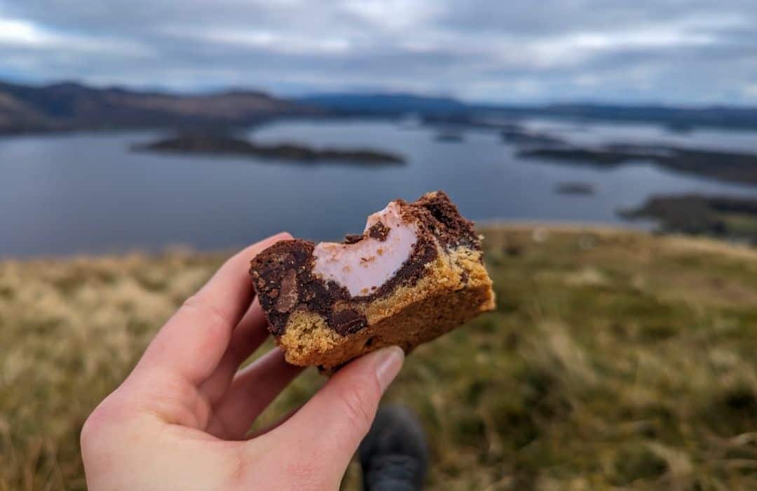 Image of a hand holding a brownie with marshmallow and biscuit with a grass mound and Loch Lomond blurred in the background. A picnic with a view. 