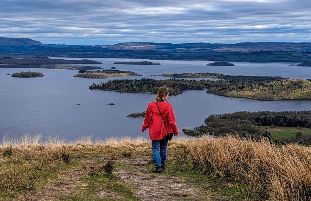 The image shows a person in a bright red jacket walking on the grassy hillside of Beinn Dubh, overlooking a scenic view of Loch Lomond, Scotland. The lake is dotted with small islands, and the surrounding landscape is a mix of rolling hills and distant mountains under a cloudy sky. The scene evokes a sense of tranquility and wide open space. It's easy to do on a Loch Lomond day trip itinerary. 