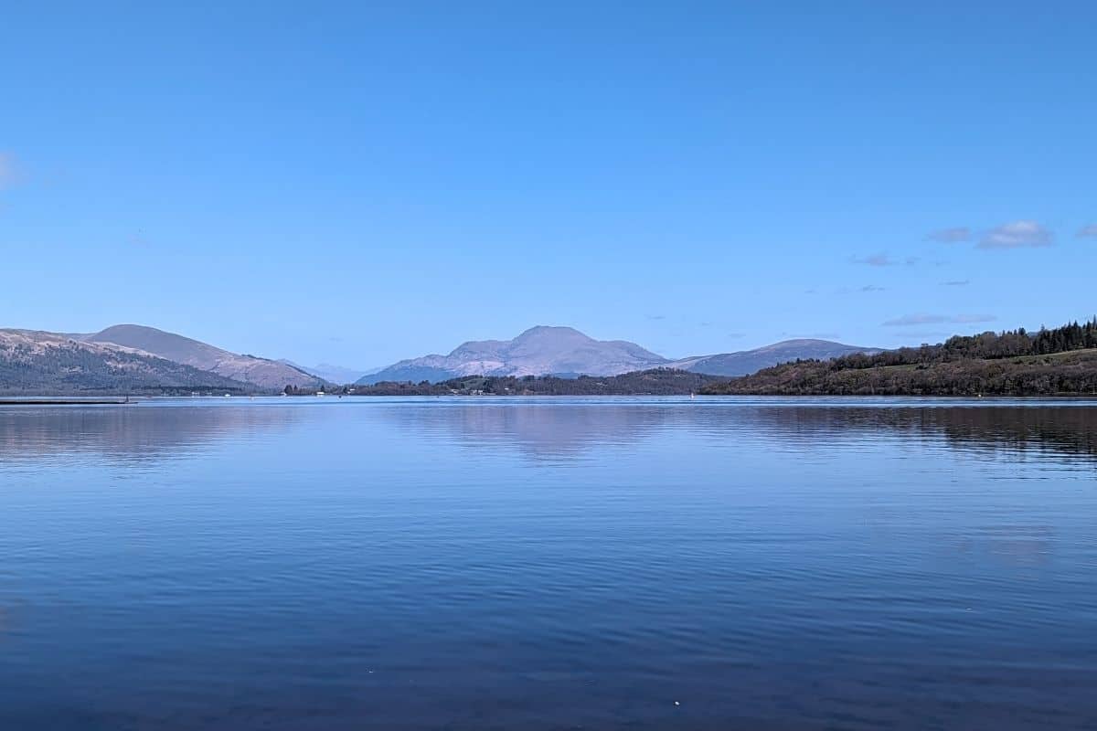 View of a loch with the blue sky reflected in the water and the rugged shape of Ben Lomond in the distance. 