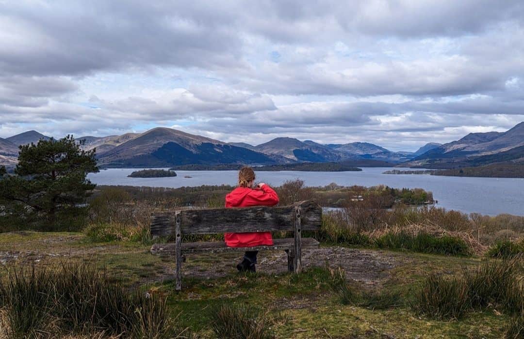 A girl in a bright red jacket sits on a wooden bench looking at Loch Lomond dotted with islands surrounded by mountains. The summit of Inchcailloch which you can visit on a Loch Lomond day trip itinerary. 