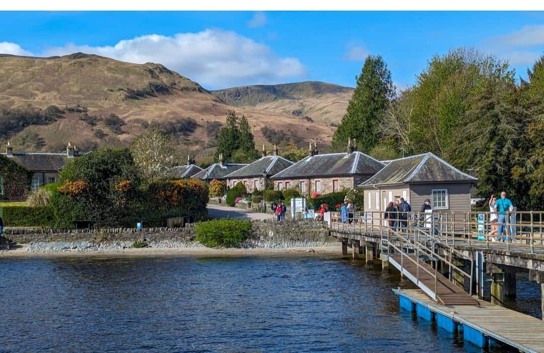 The image shows a quaint village by the edge of Loch Lomond, with traditional stone cottages lined along a quiet street. A group of people is gathered on a wooden pier that extends over the water. The backdrop features rolling hills with a mix of green and brown hues, bathed in sunlight under a partly cloudy sky. The peaceful setting captures the charm of the village of Luss.
