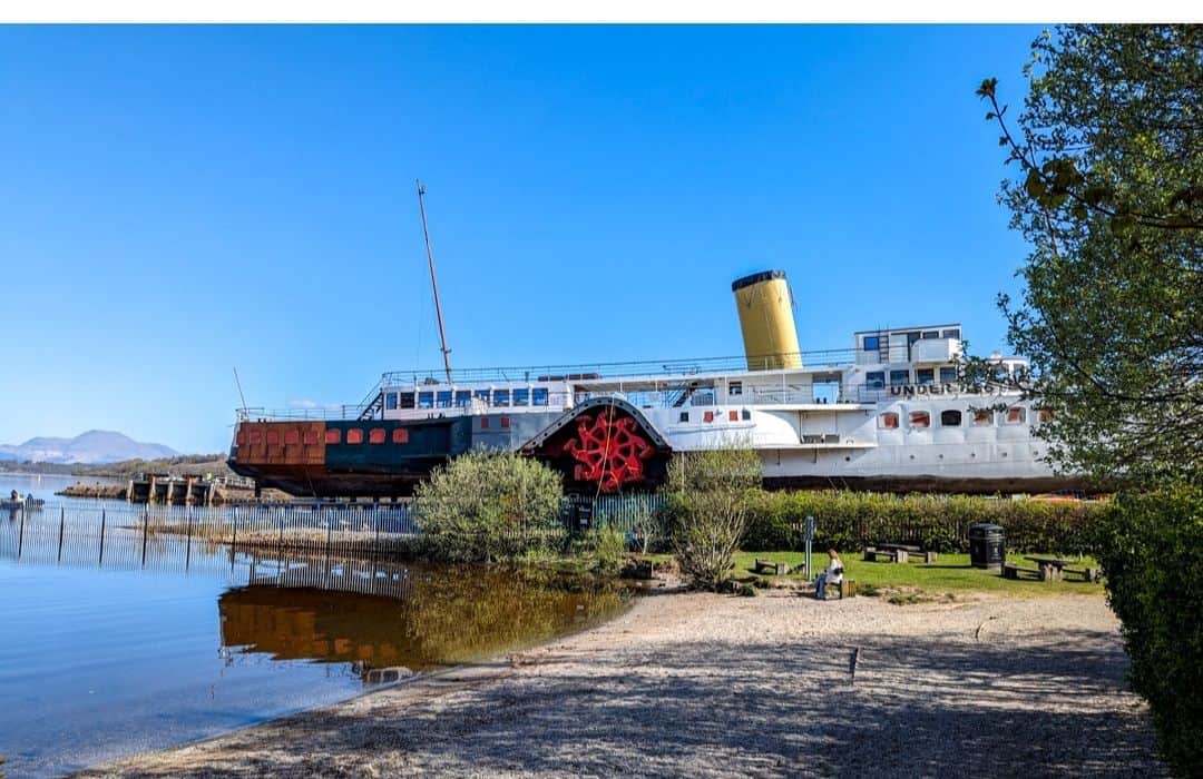 The image shows the "Maid of the Loch," a large, historic paddle steamer docked by the shore of Loch Lomond, Scotland. The steamer features a prominent yellow funnel, red paddle wheel, and white hull, with the word "UNDER" partially visible on its side. The sky is clear and blue, and the peaceful waterfront setting includes nearby greenery and a small sandy area.