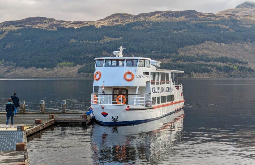 Two people walk down towards a pier where a white boat which orange paintwork waits on the waters of Loch Lomond. A rugged tree-covered hill rises up behind. 