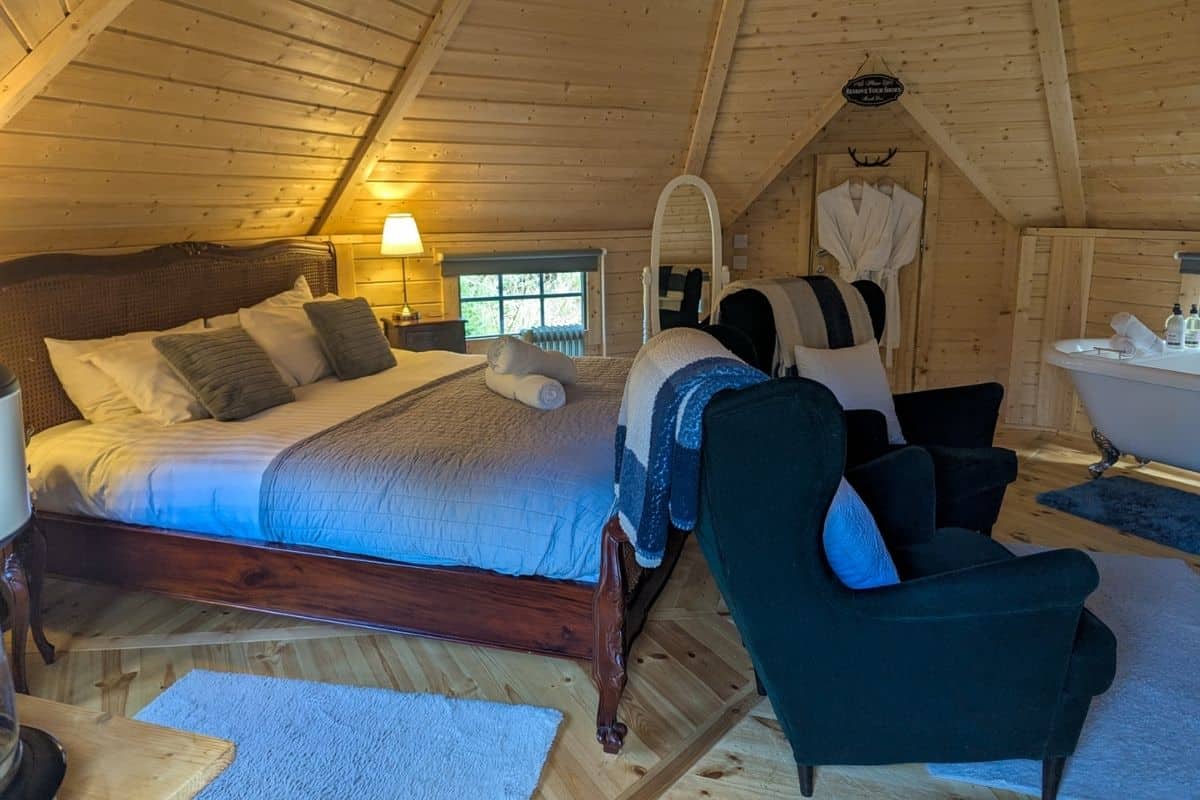 The interior of a cosy lodge with large armchairs, a double bed and a bathtub. 