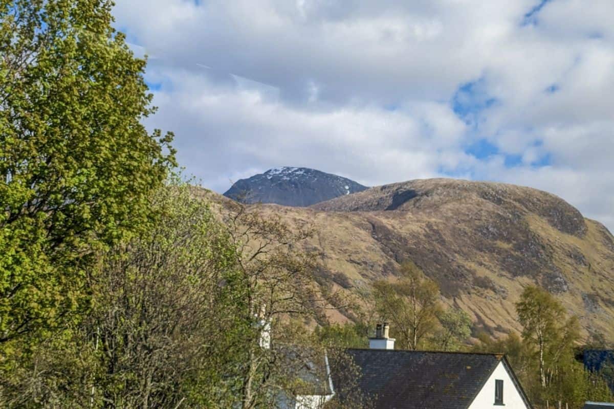 View of a steep peak rising above a huge mountainous landscape. In the front is the roof of a white house and a tree. Ben Nevis in Scotland. 