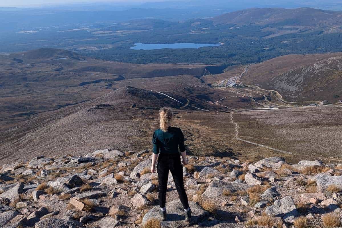 Viewing looking down at a woman looking down a rugged rocky peak towards a loch in the distance. The top of Cairn Gorm, a Munro. 