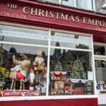 The Christmas Emporium in Pitlochry has windows filled with twinkling lights, and the entrance is framed by wreaths and garlands, creating a welcoming holiday atmosphere.