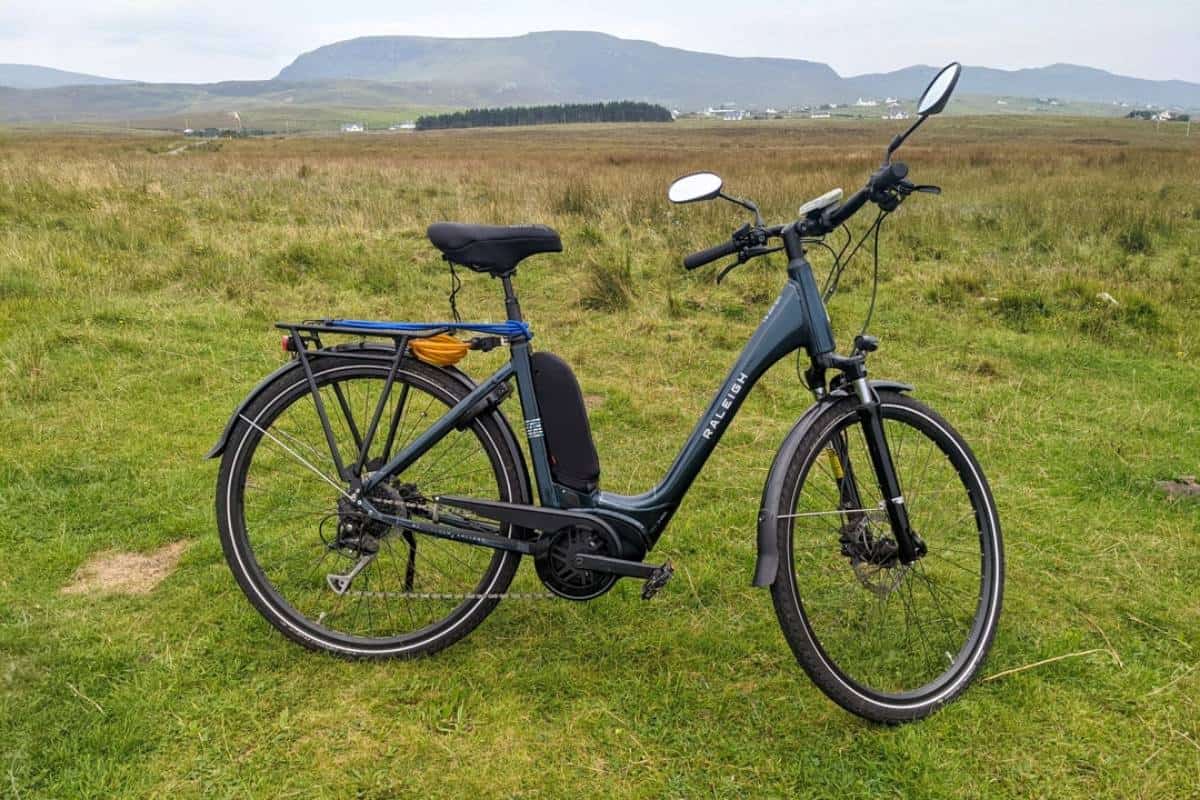 Photo of a dark green e-bike with wing mirrors standing up on grass with hills in the background. E-biking on the Trotternish Peninsula, Isle of Skye. 