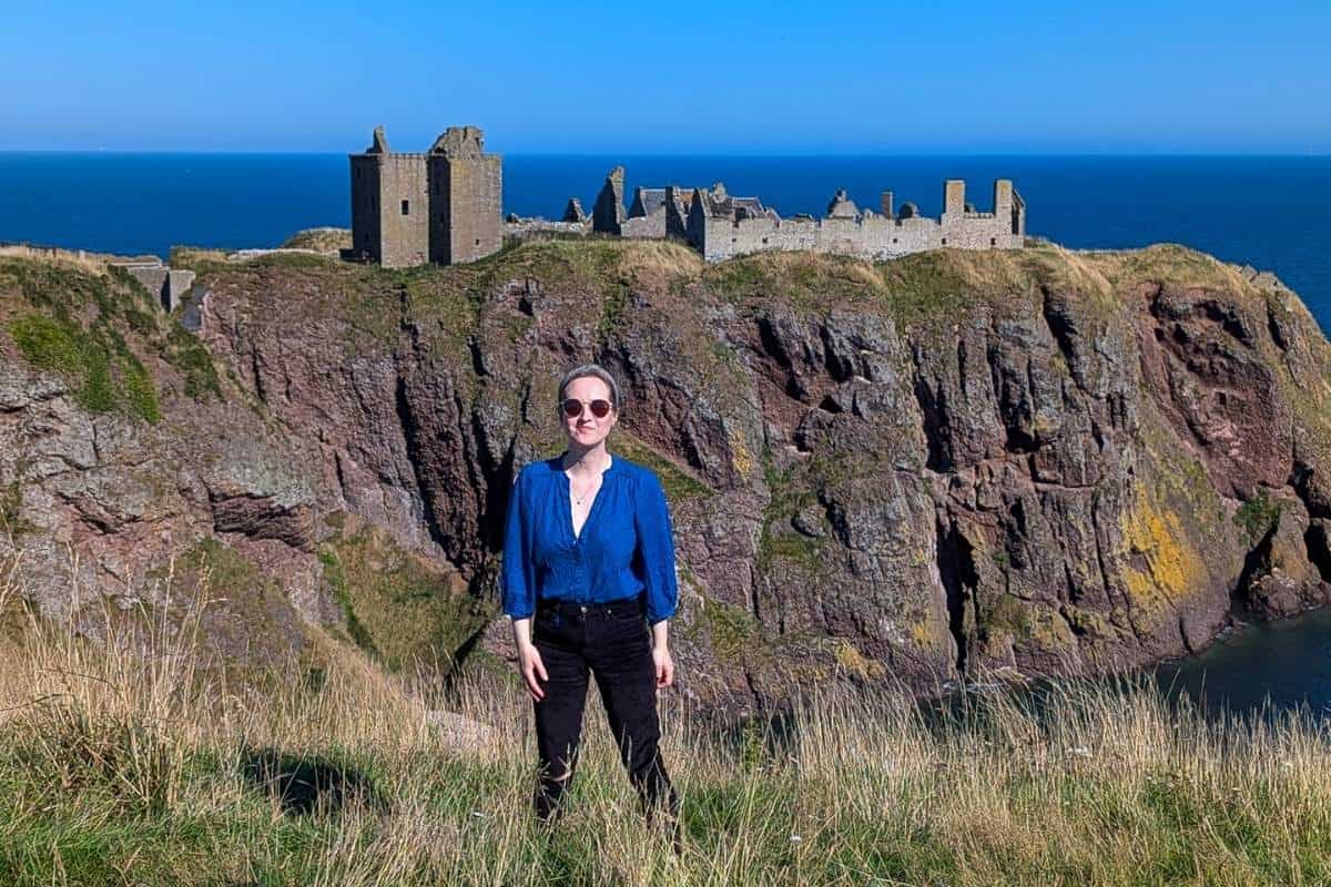 A woman wearing sunglasses and a bright blue top and black trousers stands facing the camera with the ruins of a castle on a steep crag behind her. The sea is in the background and the sky is cloudless. 