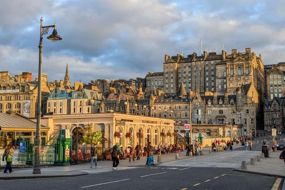 A photograph captures a bustling street scene in Edinburgh, Scotland, during the golden hour. The warm sunlight highlights the historic architecture, including grand stone buildings with intricate details and turrets. People walk along the street, some carrying luggage, while others sit on benches near a cream-colored building adorned with hanging flower baskets. A street lamp in the foreground adds a classic touch, and the sky is a mix of blue and clouds.