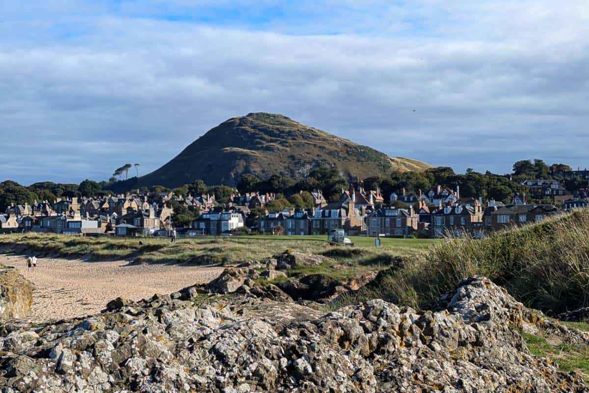 View of a steep grassy mound rising above houses. A golf course and beach are in the foreground. North Berwick. 