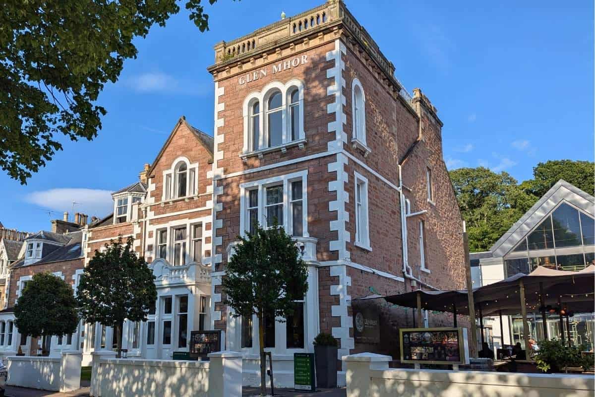 A large brick Victorian townhouse with white details and trees lining the front. A bar with a beer garden next to it. Glen Mhor Hotel in Inverness, one of the best places to stay.