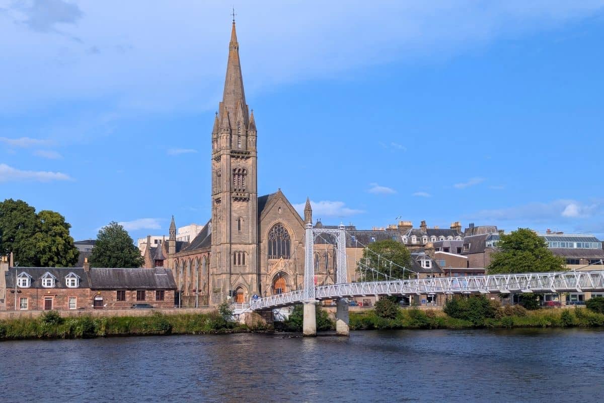 Church with a white footbridge in front of it crossing the river in Inverness on a sunny day for where to stay in Inverness. 