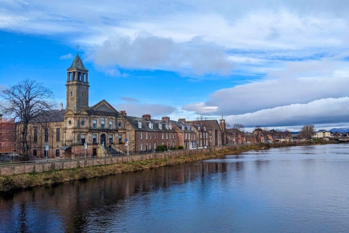 A view of the wide River Ness with Victorian buildings lining the bank. The sky is blue but there is some dark grey cloud. This is Inverness. 