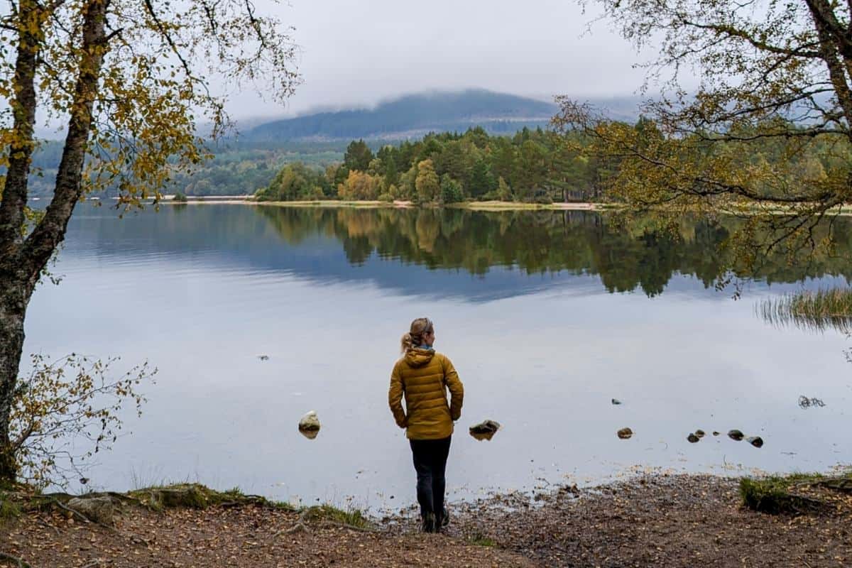 The Perfect 10-Day Scotland Itinerary by Train with Epic Views 7 A person in a yellow jacket stands at the edge of calm Loch Morlich, gazing across the still water that mirrors the surrounding forest and mist-covered mountain. Autumn leaves frame the serene scene, capturing a peaceful moment in the Scottish Highlands near Aviemore.