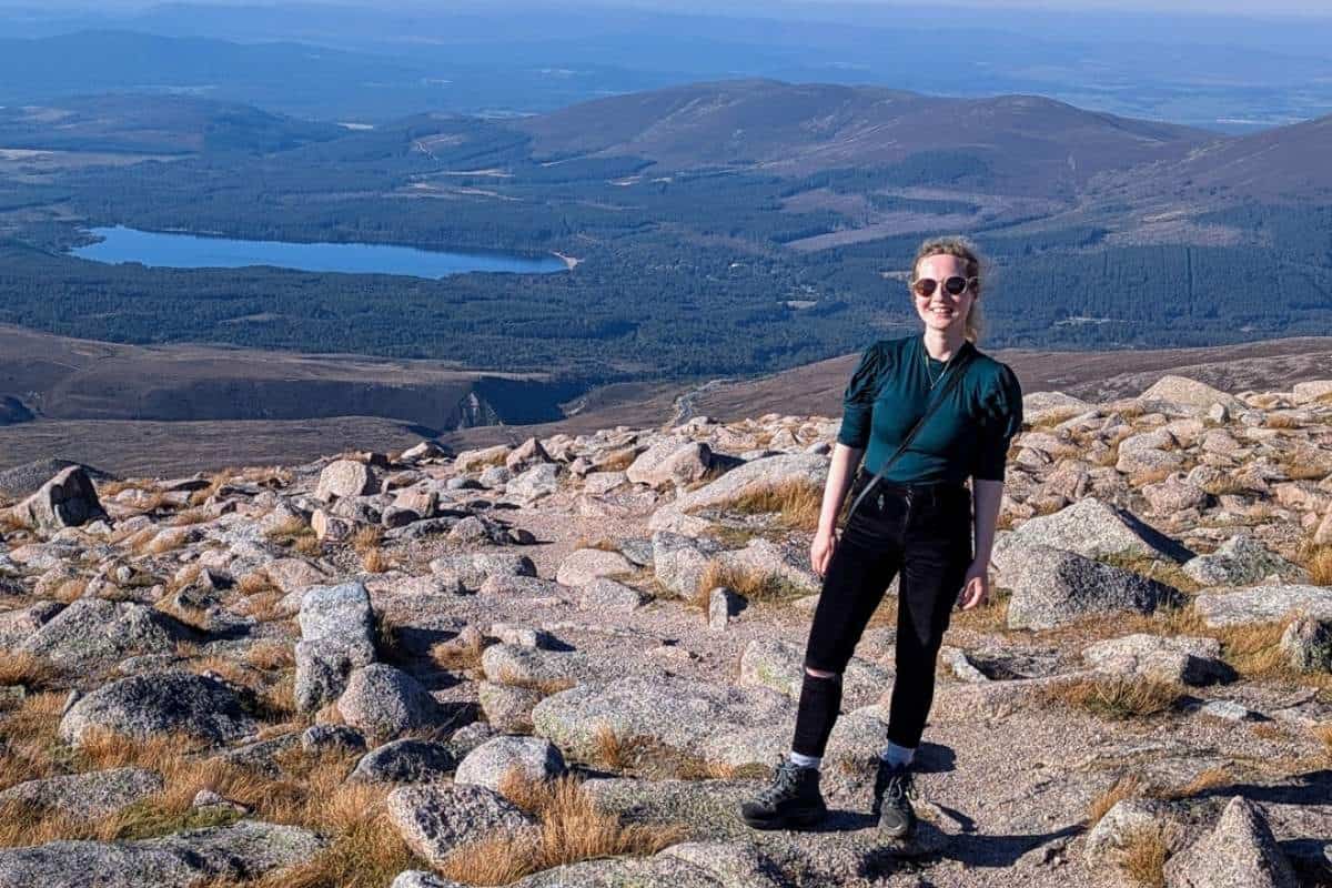 Best Time to Visit Scotland (& The Worst!) 10 A woman wearing a green top and black trousers and sunglasses stands on a mountain top looking down onto a loch and forest. It's sunny. September is the best time to visit Scotland for sunshine without the summer crowds.