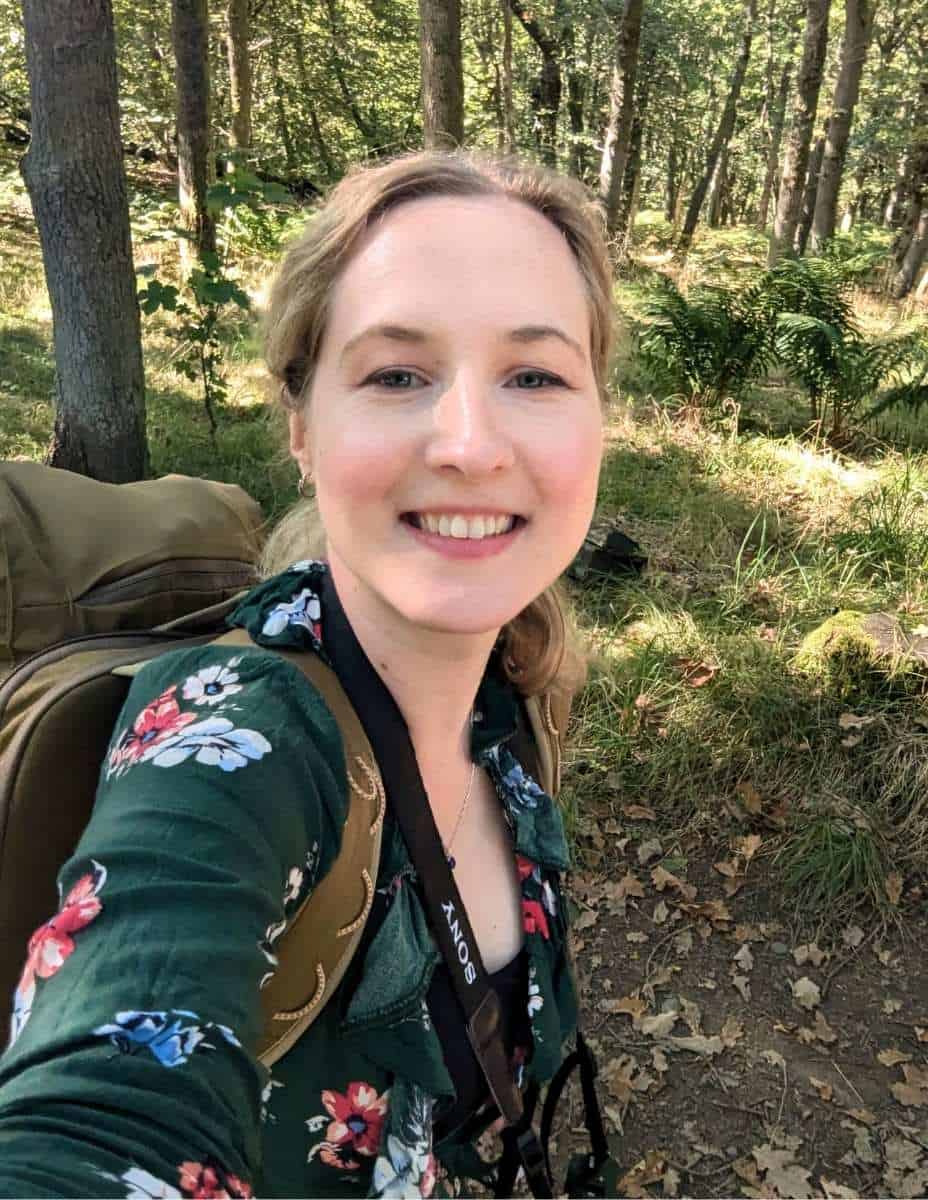 A woman smiling for a selfie in a sunlit forest, wearing a green floral top and carrying a large khaki backpack with a camera strap around her neck. She is Francesca, the founder of Little Lost Travel and the expert behind 'How to Plan a Trip to Scotland.'