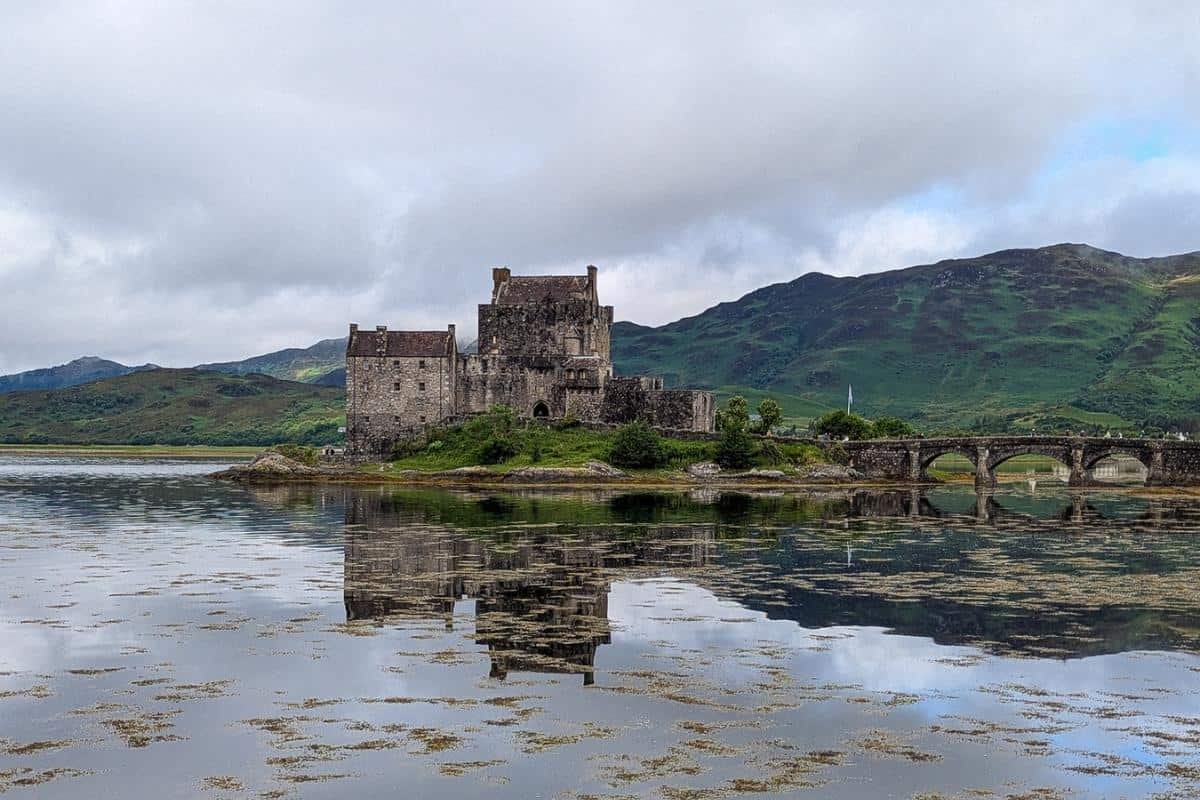 Best Time to Visit Scotland (& The Worst!) 7 The photo shows Eilean Donan Castle perched on a small tidal island where three sea lochs meet in the Scottish Highlands. The stone castle, connected to the mainland by an arched stone bridge, is reflected in the calm water, with lush green hills rising in the background under a cloudy sky. May is the best time to visit Scotland as the landscape is lush and green.