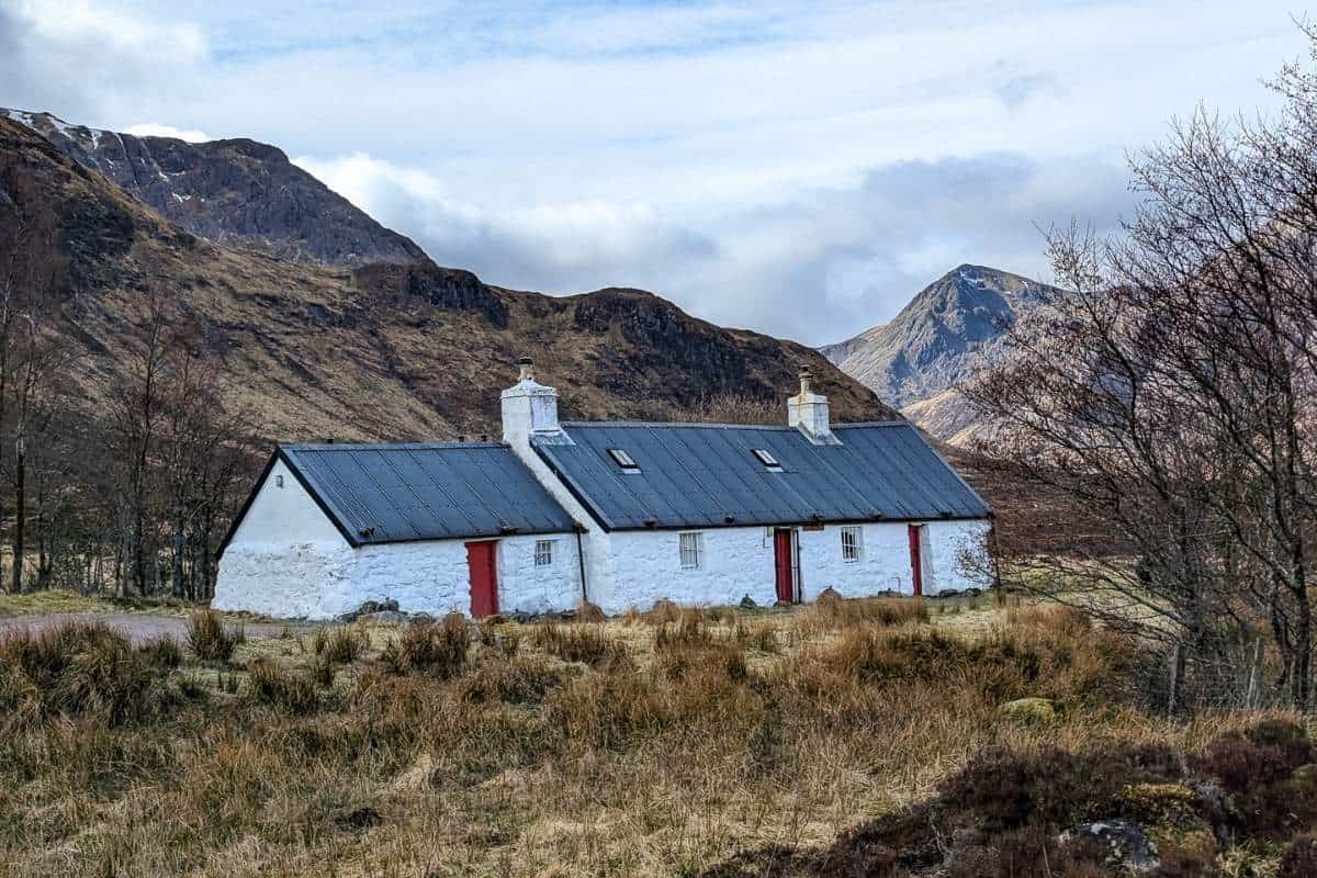 Best Time to Visit Scotland (& The Worst!) 4 This image shows a traditional white stone cottage with a slate roof and red doors set against the dramatic, rugged hills of the Scottish Highlands under a cloudy sky. The surrounding landscape features brown heather and sparse trees, evoking a remote and peaceful atmosphere.