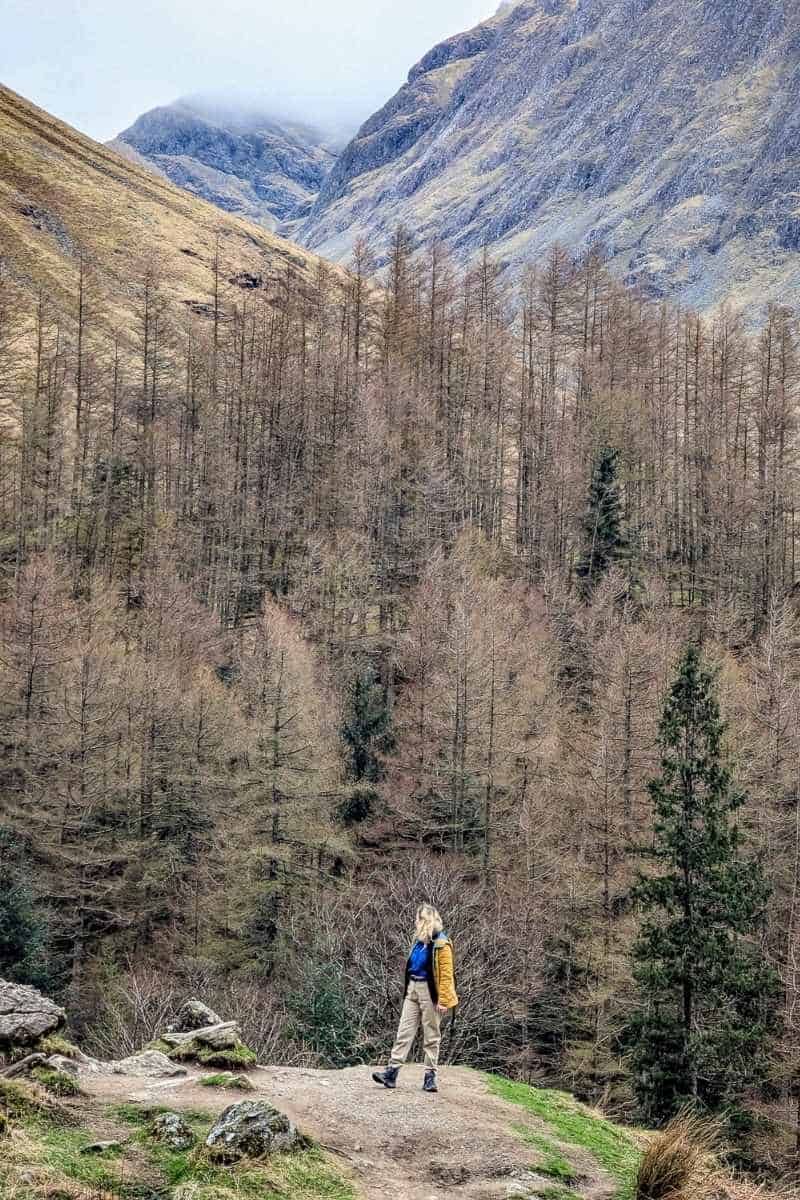 Best Time to Visit Scotland (& The Worst!) 2 A woman wearing a yellow jacket and beige trousers stands on a ledge. Behind her is a brown forest in the winter with misty mountains behind. Hagrid's Hut in Glencoe in winter.