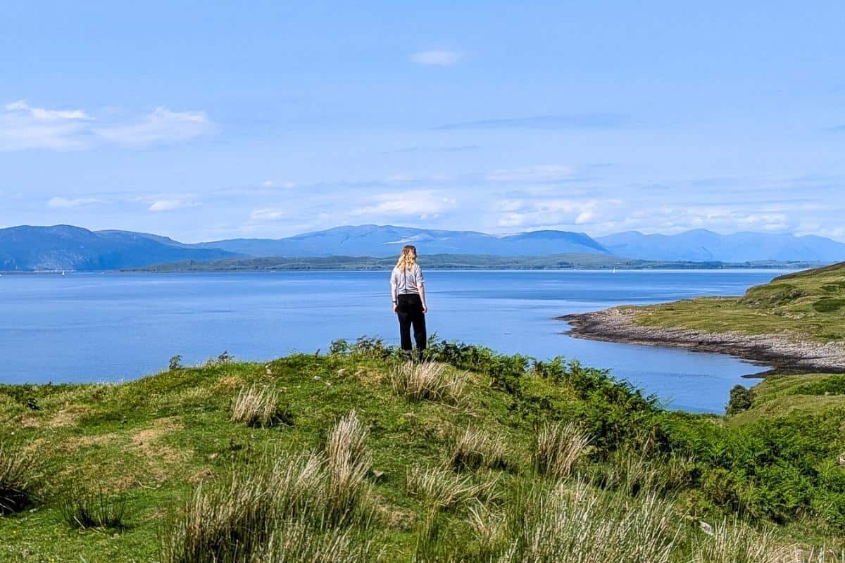 Best Time to Visit Scotland (& The Worst!) 1 A woman wearing black trousers and a white t-shirts stands on a green grassy moundoverlooking the Sound of Mull with mountains in the distance. The sky is blue wth some white cloud. May is the best time to visit Scotland for drier weather on average.