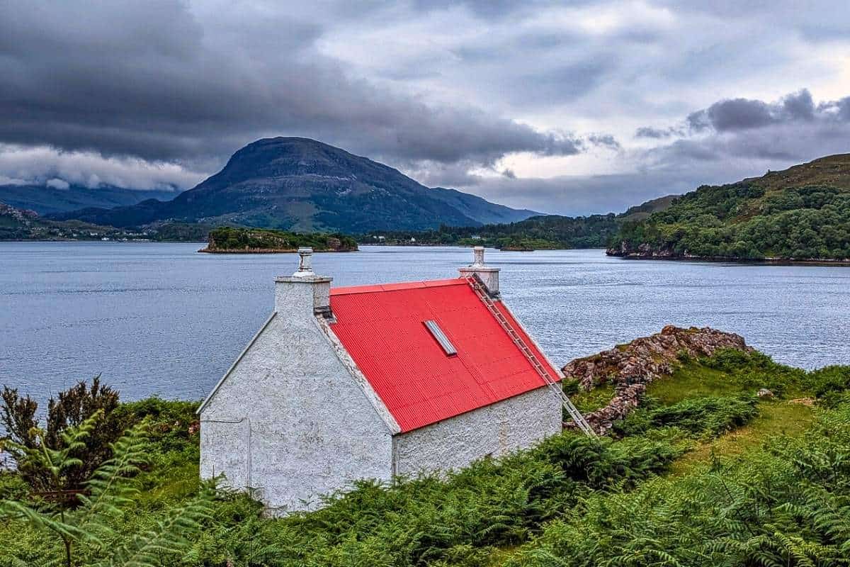 Best Time to Visit Scotland (& The Worst!) 9 A white cottage with a red roof and a white ladder going up the side its on a grassy shoreline overlooking a large loch with mountains in the distance. The sky is heavy and grey.