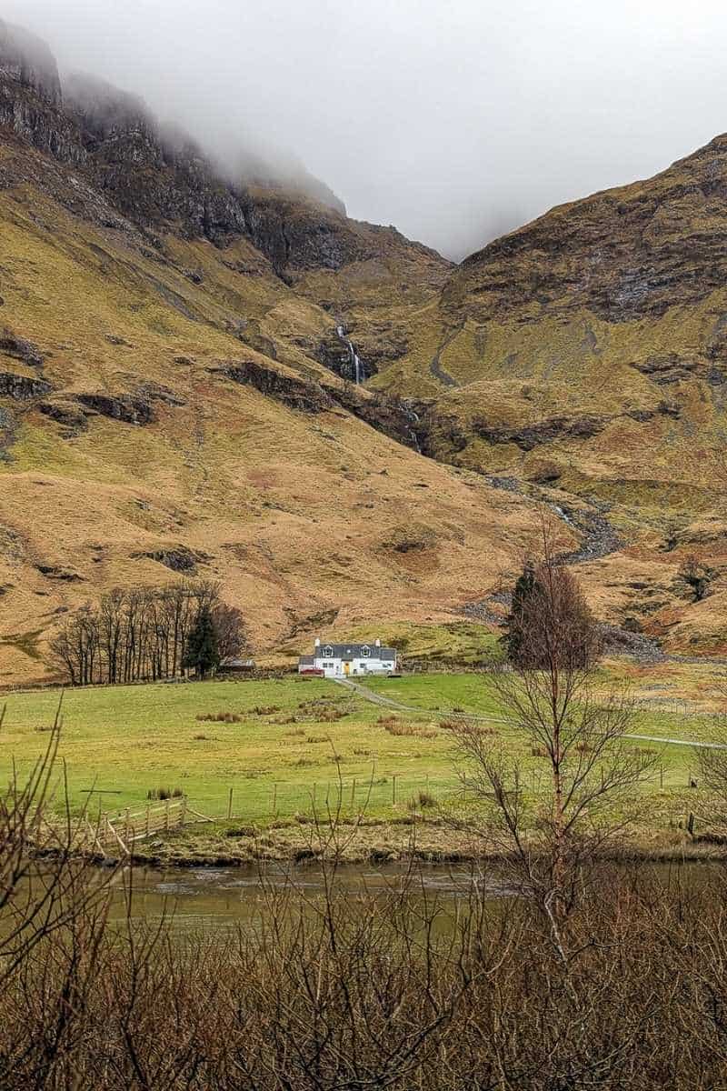 Best Time to Visit Scotland (& The Worst!) 3 A little white cottage sits with a tall mountain peak behind it. A stream runs down the side of it. This is Glencoe in winter.