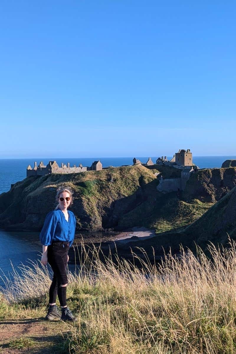13 Best Places to Visit in Scotland by Train 3 A visitor standing on grassy cliffs with castle ruins in the background by the sea, capturing the charm of the best places to visit in Scotland by train.