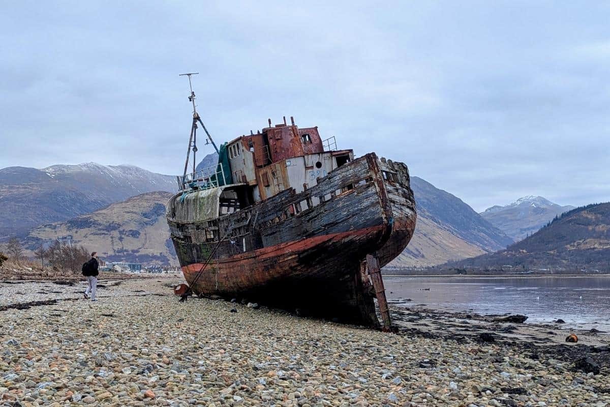 13 Best Places to Visit in Scotland by Train 8 Shipwrecked boat on a pebble beach beside a loch with snow-capped mountains behind. Corpach near Fort William.