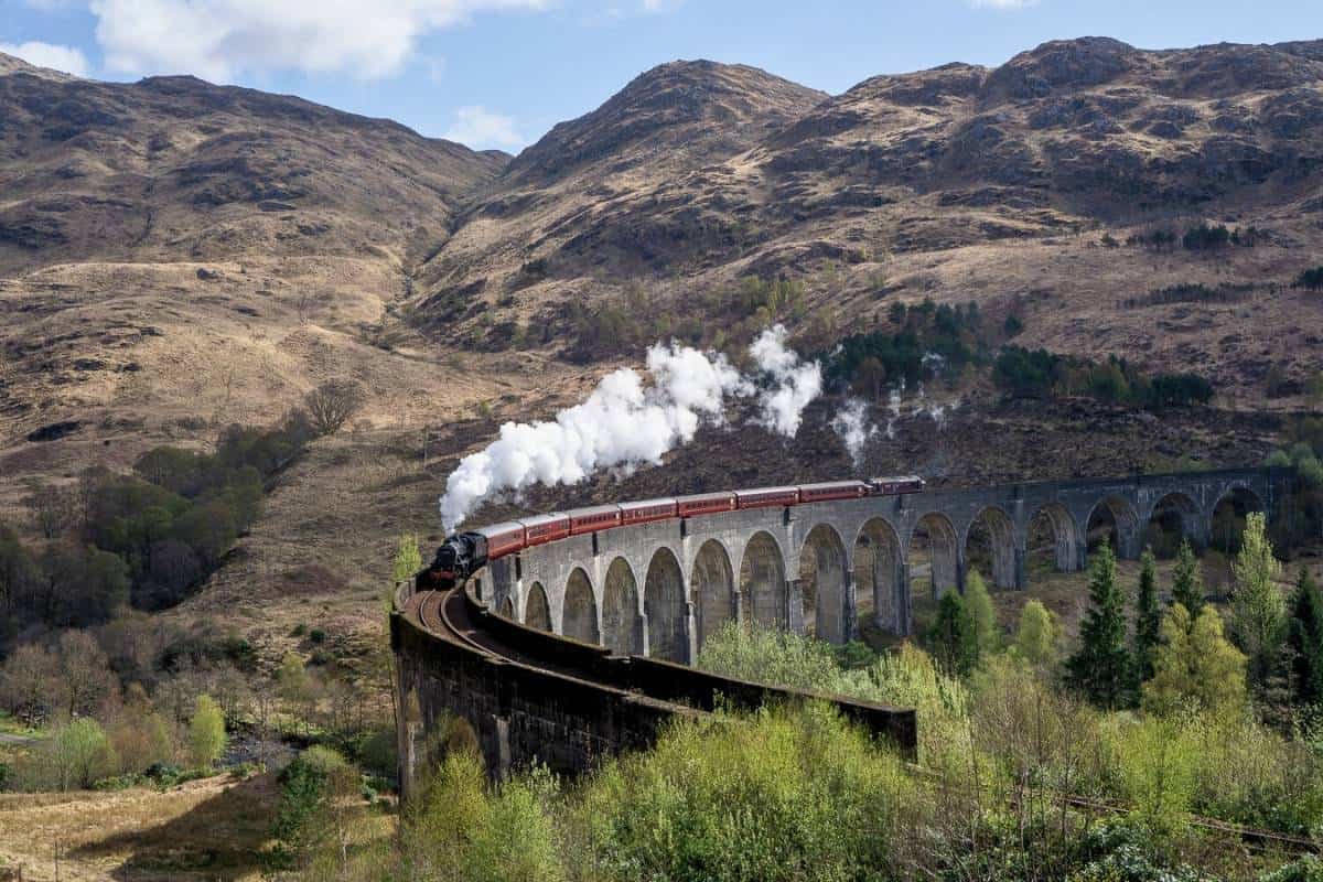 A vintage black steam locomotive with red carriages crosses the tall stone arches of the Glenfinnan Viaduct, with white smoke billowing against the backdrop of green Scottish Highlands hills.