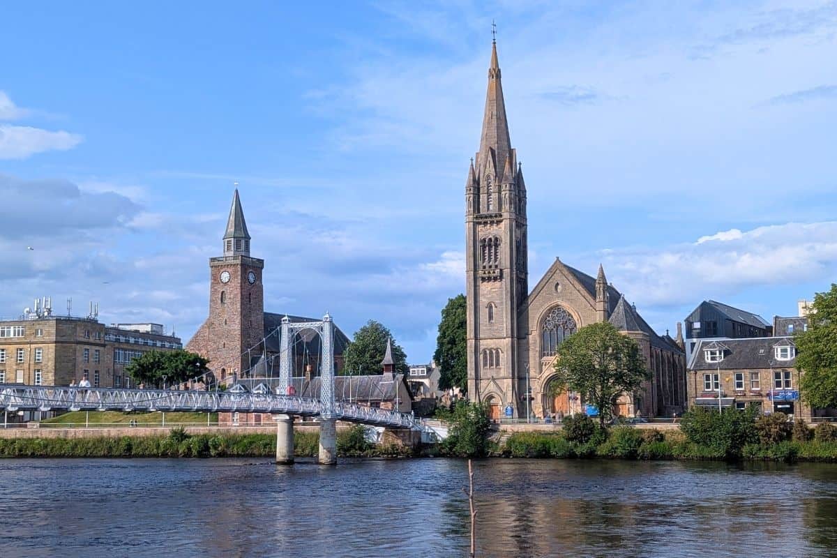 13 Best Places to Visit in Scotland by Train 4 A sunny day view of Inverness with a bridge crossing the River Ness and several churches lining the riverbank, their spires rising against the blue sky.