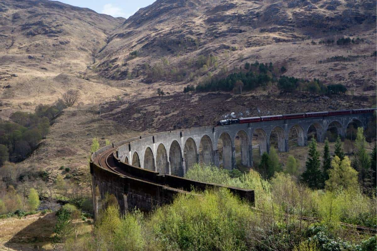 13 Best Places to Visit in Scotland by Train 16 A dark red train puffs steam as it stars to cross a curved viaduct surrounded by trees and mountains.