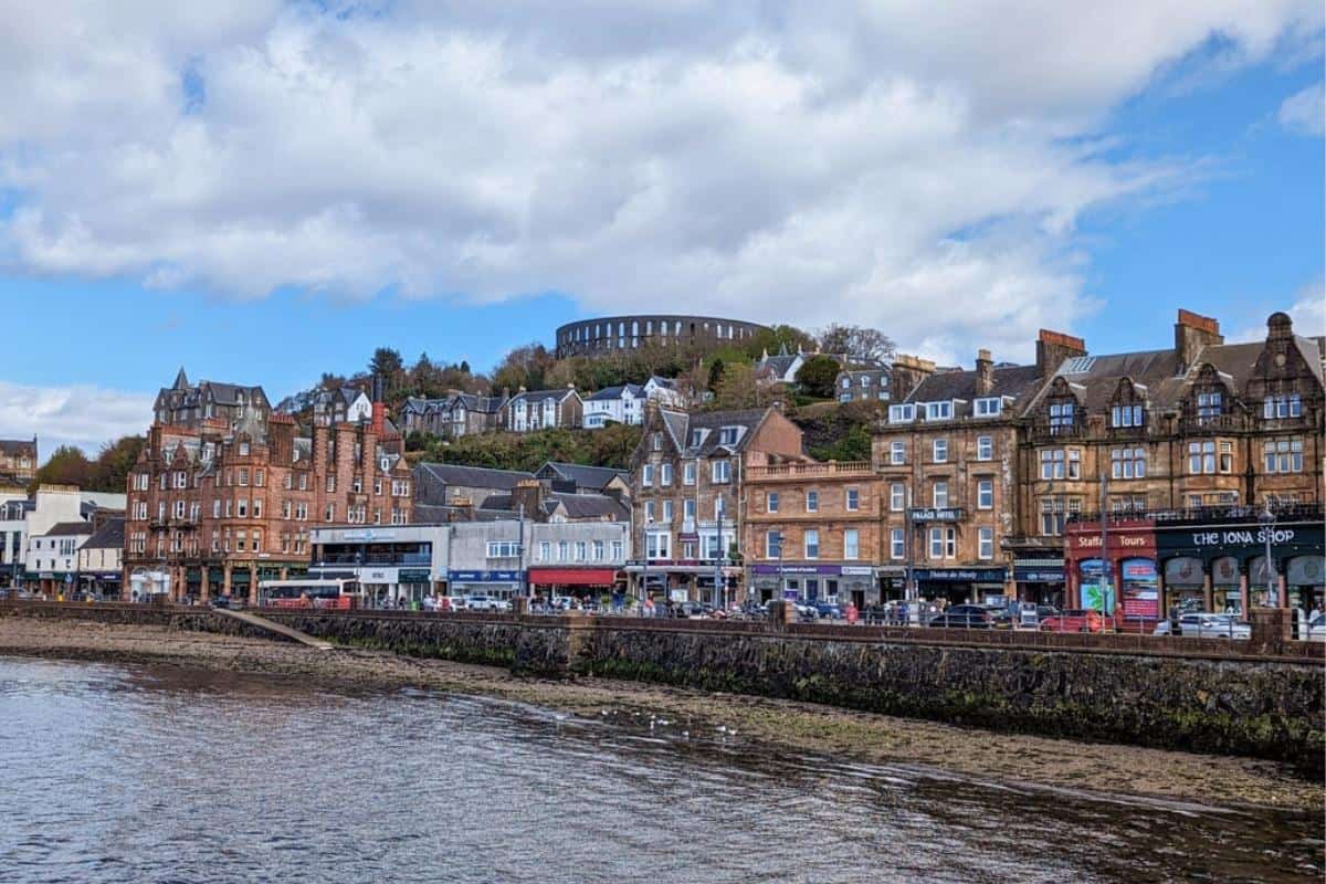 13 Best Places to Visit in Scotland by Train 10 Oban promenade lined with shops. Behind it on the hill surrounded by trees is a round tower. Oban is one of the best places to visit in Scotland by train.