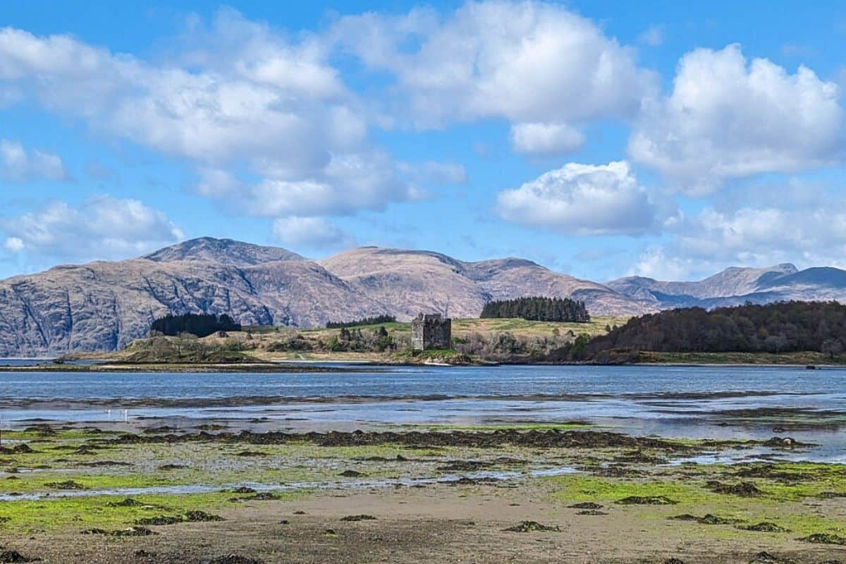 View of Castle Stalker in Scotland from acoss the water. A fortress castle sits on an island with mountains behind. One of the castles on this Rabbie's tour. 