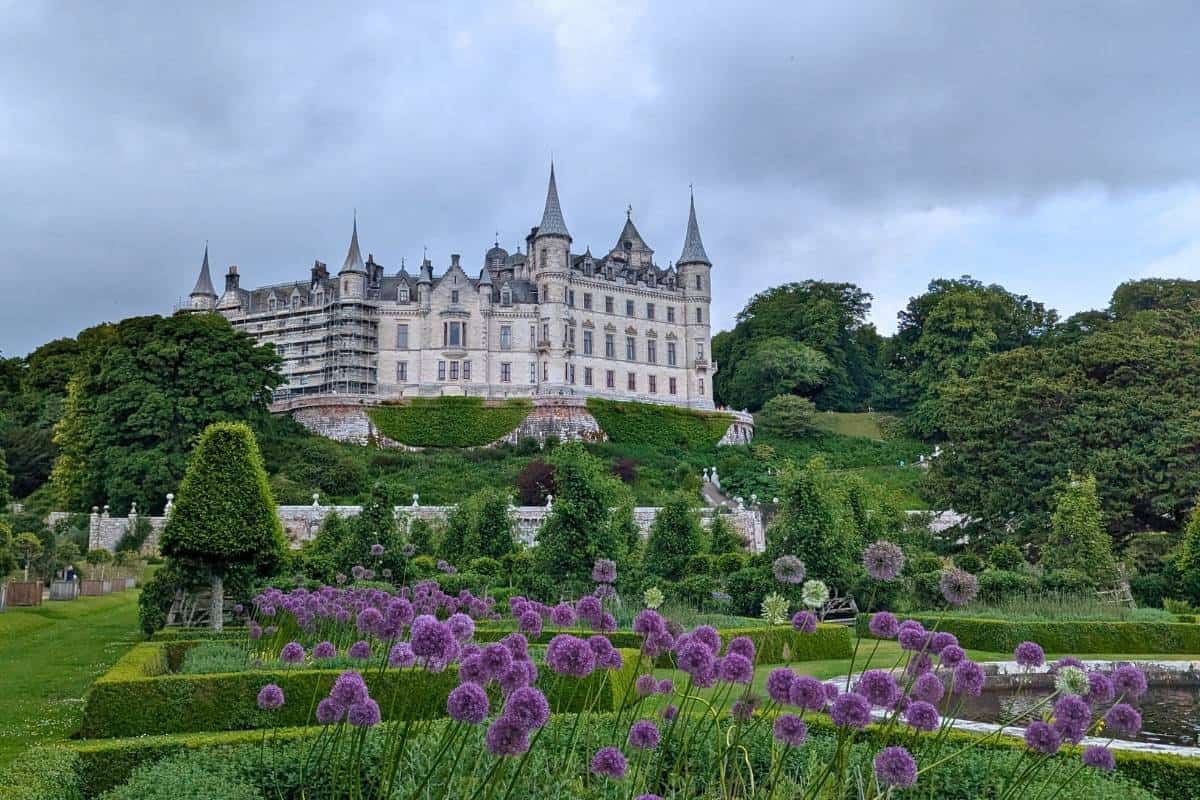 A dramatic view of Dunrobin Castle in Scotland, perched on a lush green hillside with sharp spires rising against a moody, overcast sky. In the foreground, a circular fountain sprays water into the air, surrounded by manicured gardens and topiary shrubs.