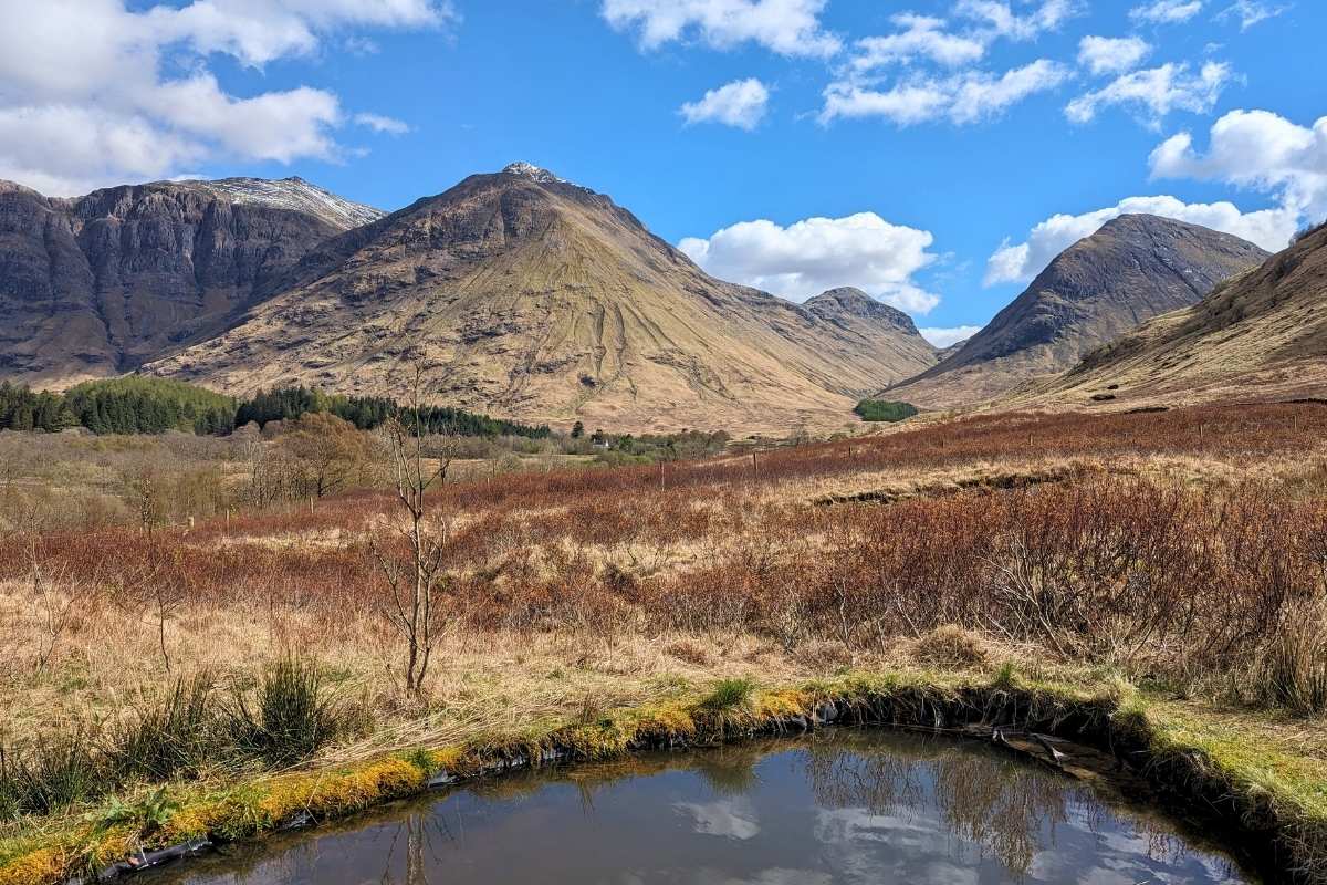 Rugged brown mountain peaks with a pond in the front. Blue sky above. Glen Coe fom the visitor centre on this Rabbie's tours review. 