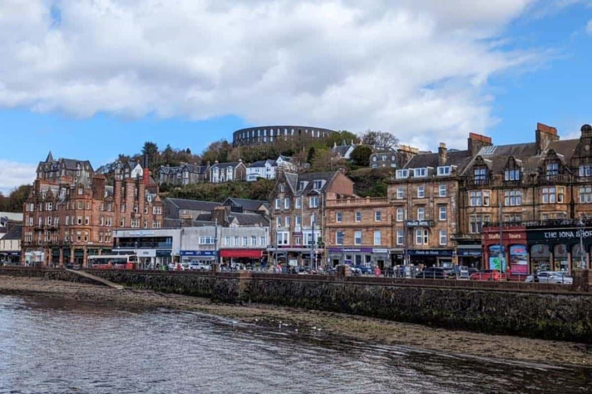 View from across the water of Oban. Houses and shopfronts line a street with a round tower on the hill above them.