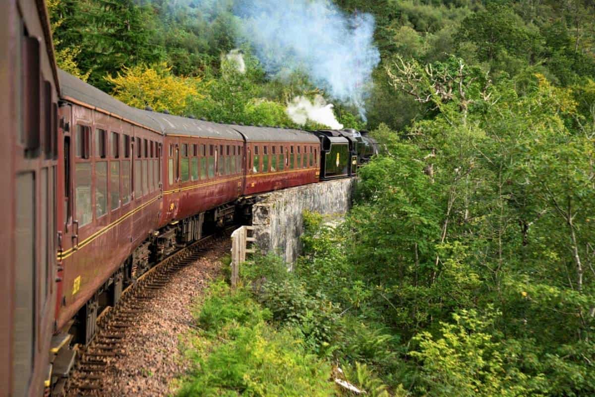 7 Most Scenic Train Journeys in Scotland with Epic Views 5 The Jacobite steam train on a track surrounded by trees and puffing steam. It's one of the most scenic train journeys in Scotland.