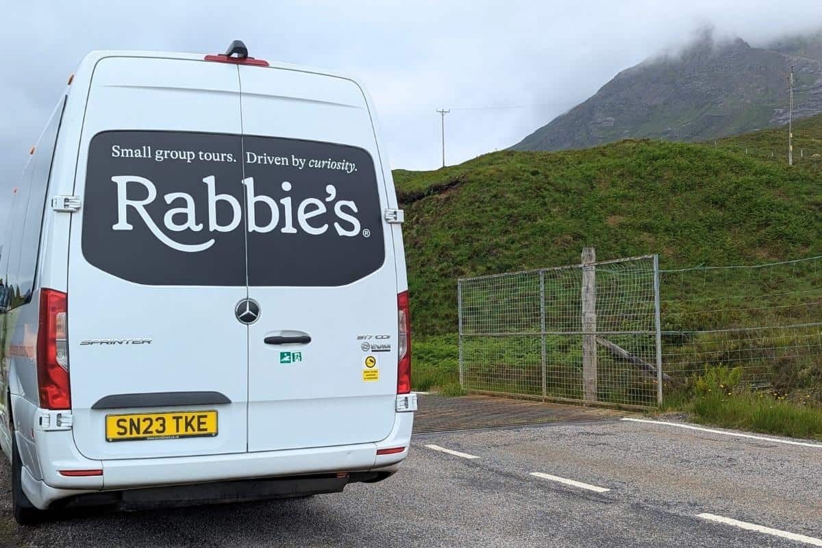 The back of a Rabbie's bus with the words 'Small group tours, driven by curiosity' on it. It's aprked at the side of a street with mountains behind.